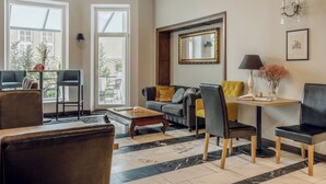 Un salon moderne comprenant un canapé, des fauteuils, une table basse et une table d’appoint. On y trouve de grandes fenêtres, un miroir et un lustre.
