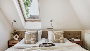 Une chambre avec une lucarne, un lit agrémenté d’oreillers à motifs, une table de chevet avec un livre et un vase, et une petite table de nuit avec une lampe.