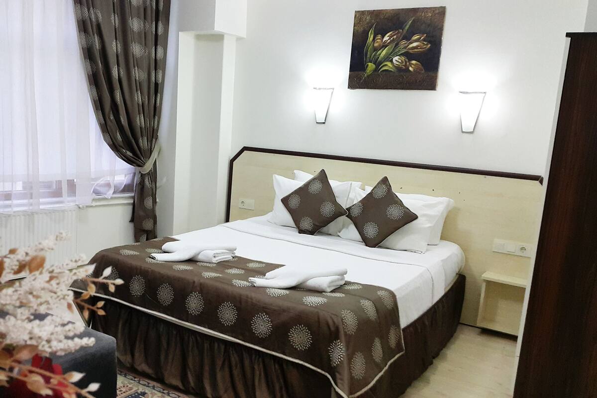 A hotel room with a bed, a nightstand, a wall-mounted lamp, a window with curtains, and a painting on the wall.