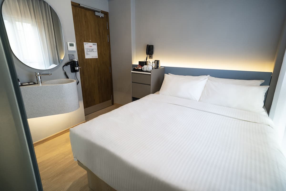 A hotel room with a bed, a bedside table, a mirror, a sink, and a phone.