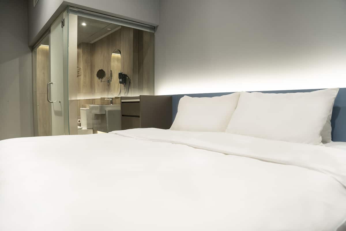 A modern hotel room with a large bed, a glass-enclosed walk-in closet, and a small bathroom with a sink and mirror.