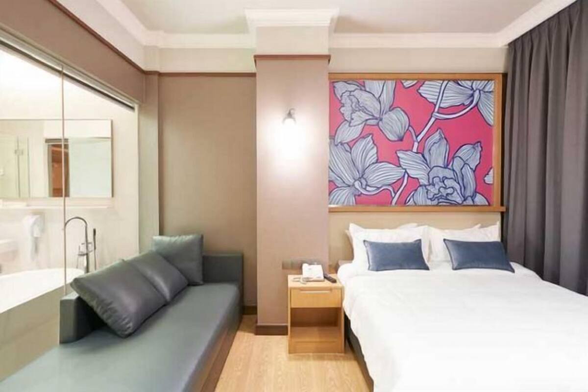 A hotel room with a bed, a sofa, a nightstand, a mirror, and a floral artwork on the wall.