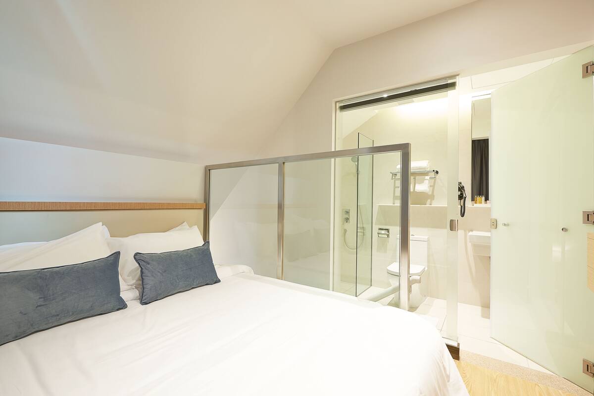 A modern hotel room with a large bed, a glass shower enclosure, and a separate bathroom visible through the glass.