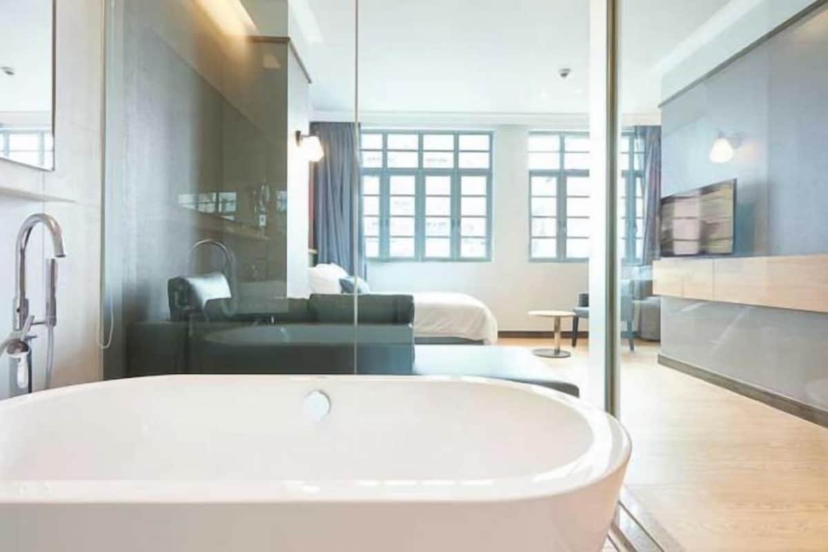 Une salle de bain moderne avec une baignoire sur pieds, une douche en verre et une chambre visible, avec un lit et une table de chevet.