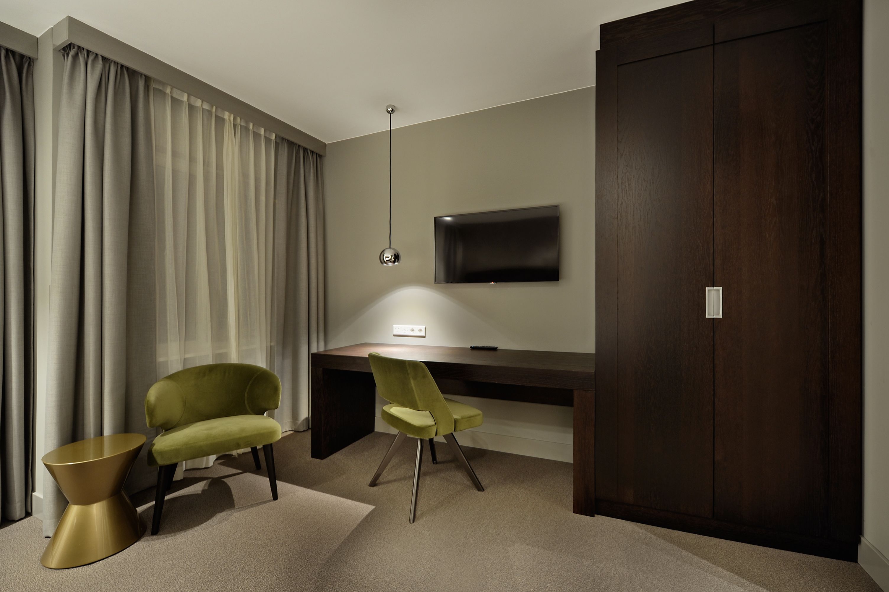 A modern hotel room with a dark wood desk, a green chair, a gold side table, and a flat-screen TV mounted on the wall.
