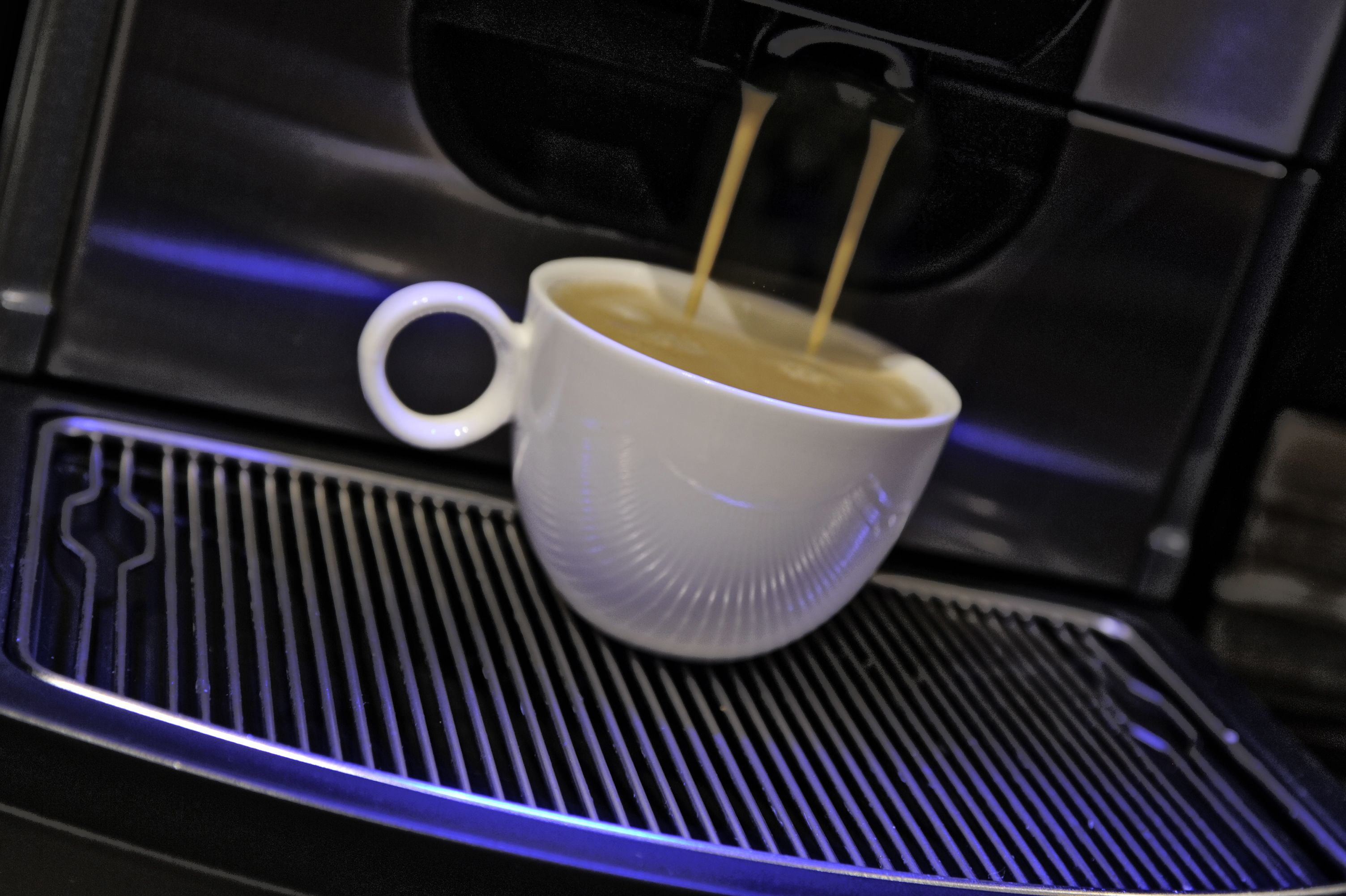 Una tassa de cafè blanca sobre una superfície metàl·lica, amb una màquina de cafè a dalt.