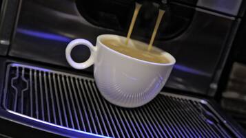 Una tassa de cafè blanca sobre una superfície metàl·lica, amb una màquina de cafè a dalt.