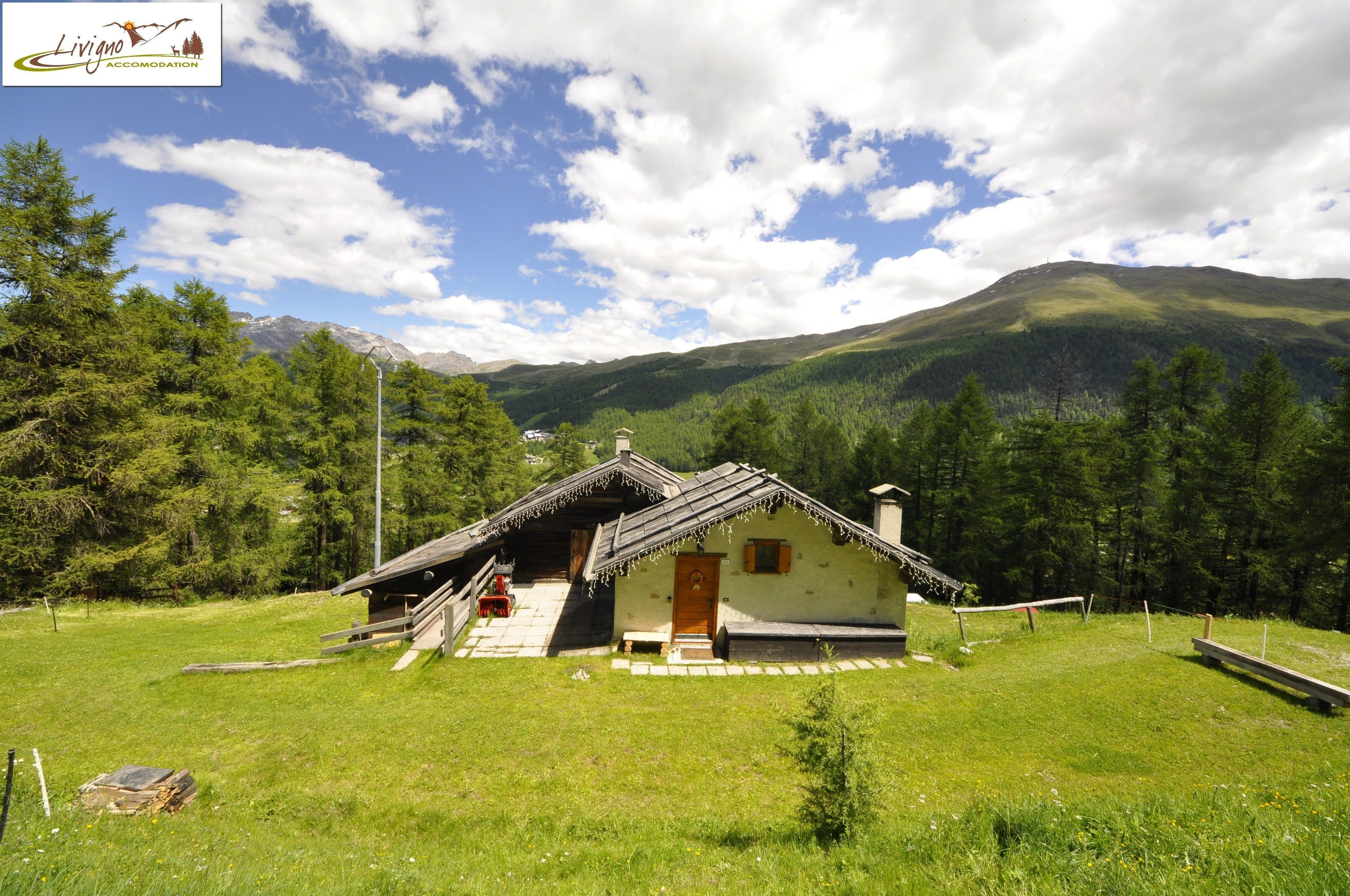 A traditional chalet with a wooden staircase leading to the entrance, surrounded by lush greenery and mountains in the background.