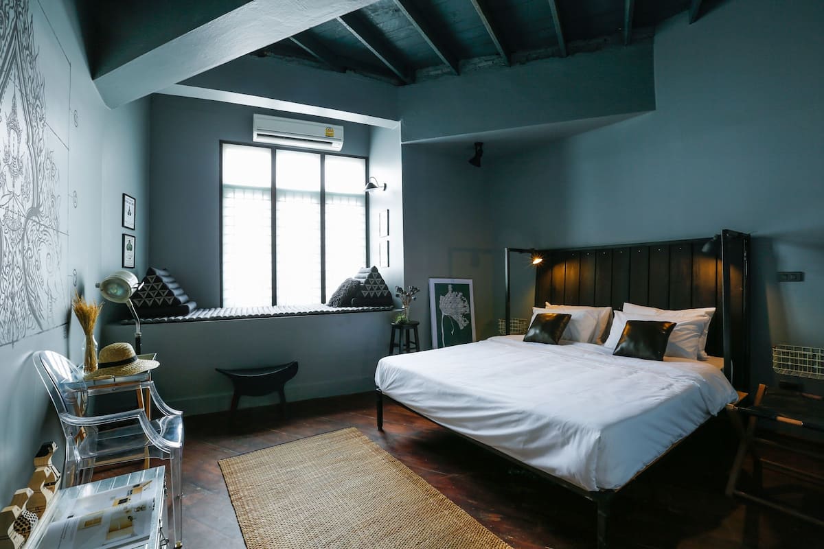 A bedroom with a large bed, a window with blinds, a desk with a chair, and a wall-mounted air conditioner.