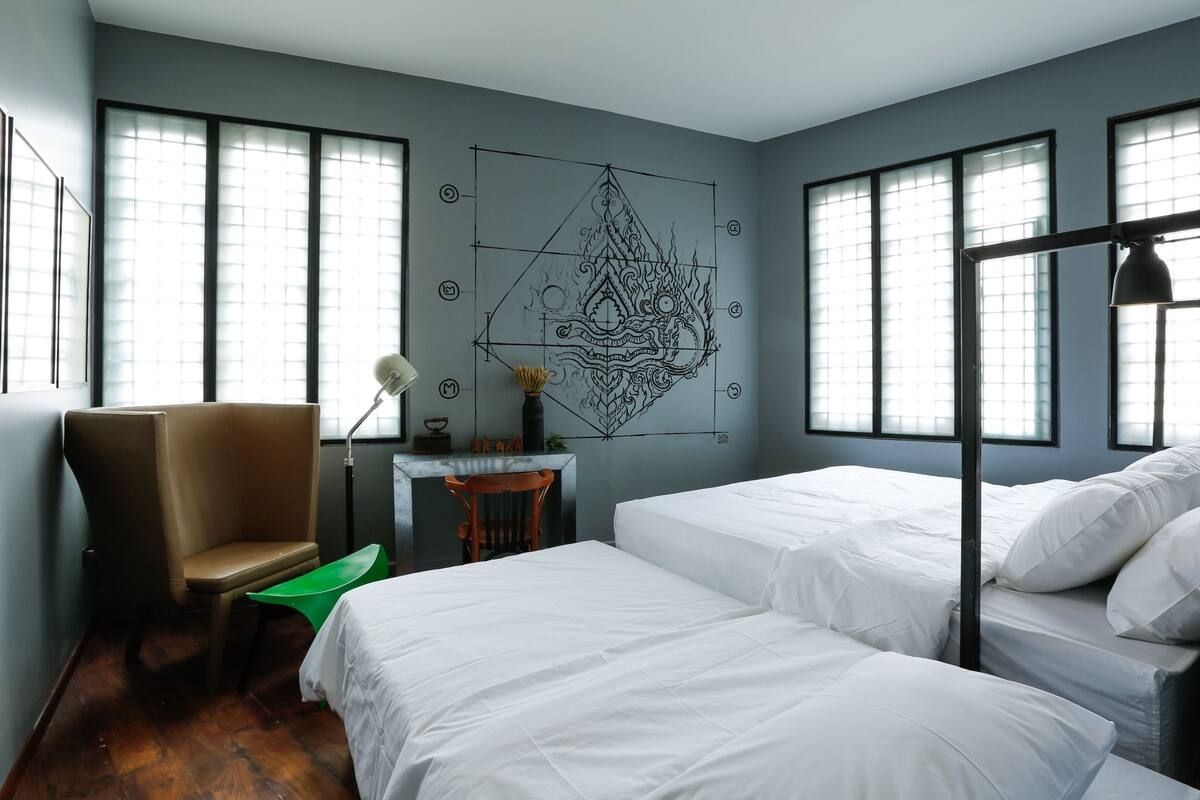 A room with two beds, a chair, a desk, and a wall art piece.
