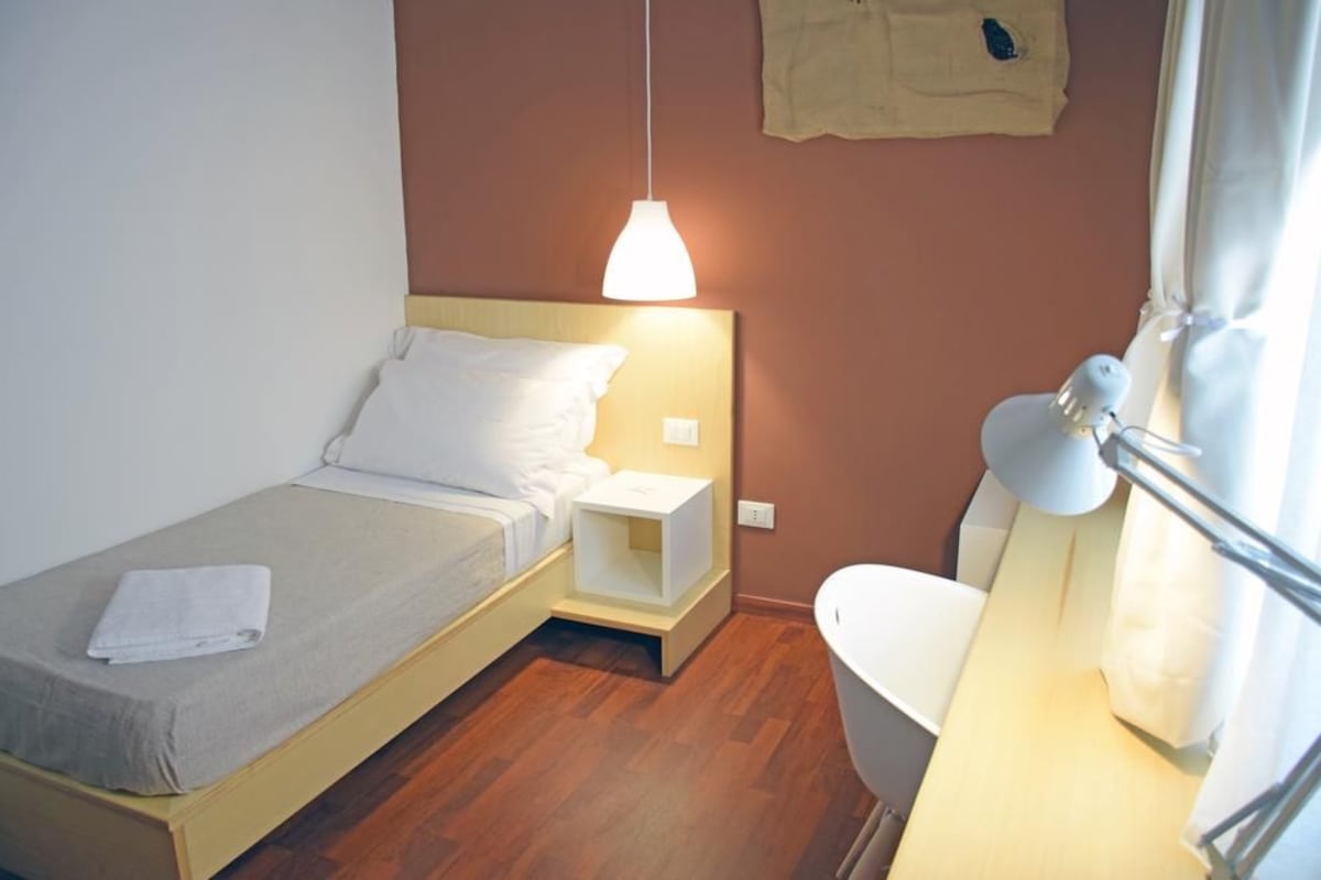 Un llit individual amb capçalera, una taula de nit, una làmpada penjant i un bany amb un vàter blanc i un penjador per tovalloles.