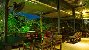 An outdoor seating area with wooden tables and chairs, surrounded by glass walls and a view of trees. - Wet Water Resort (Gampaha)
