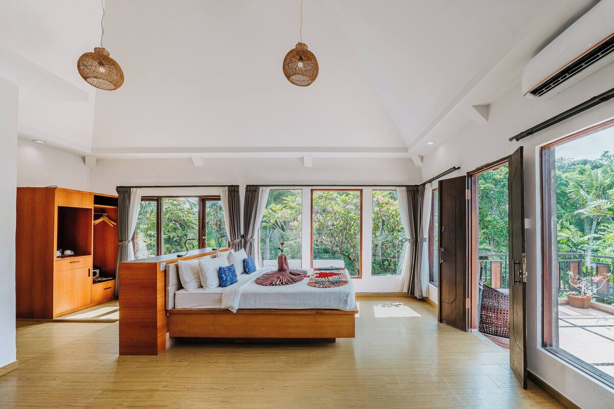 A spacious bedroom with a large bed, wooden wardrobe, and a view of greenery through large windows.