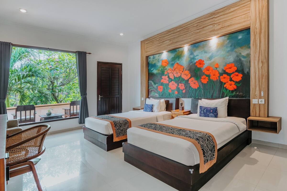 A hotel room with two beds, a large window with curtains, a painting of flowers on the wall, and a balcony with a view of greenery.