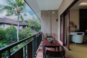 A balcony with a wooden table and chairs, a view of a garden with palm trees, and a glimpse of a kitchen area.