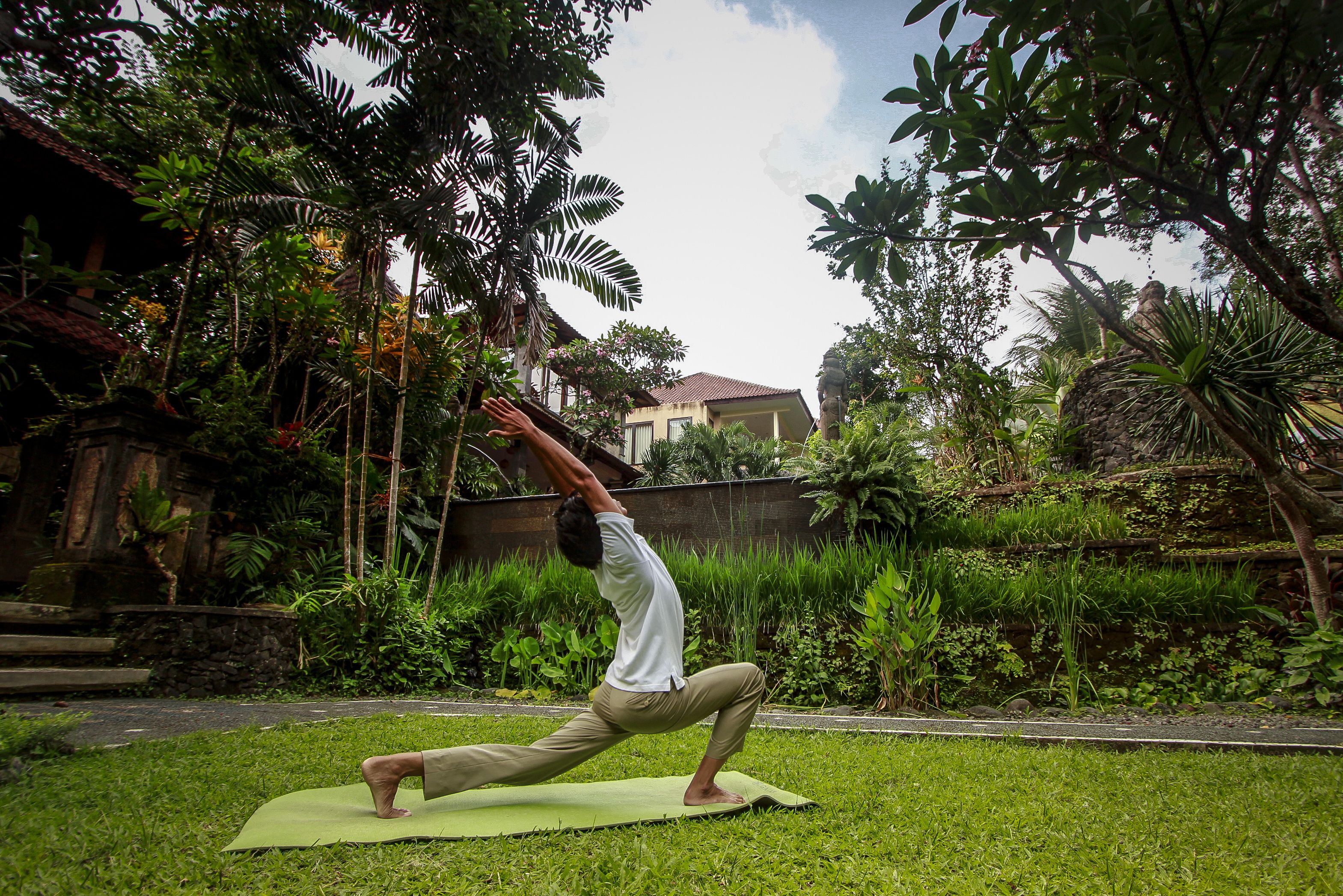 A person performing a yoga pose on a mat in a garden with lush greenery and a building in the background.