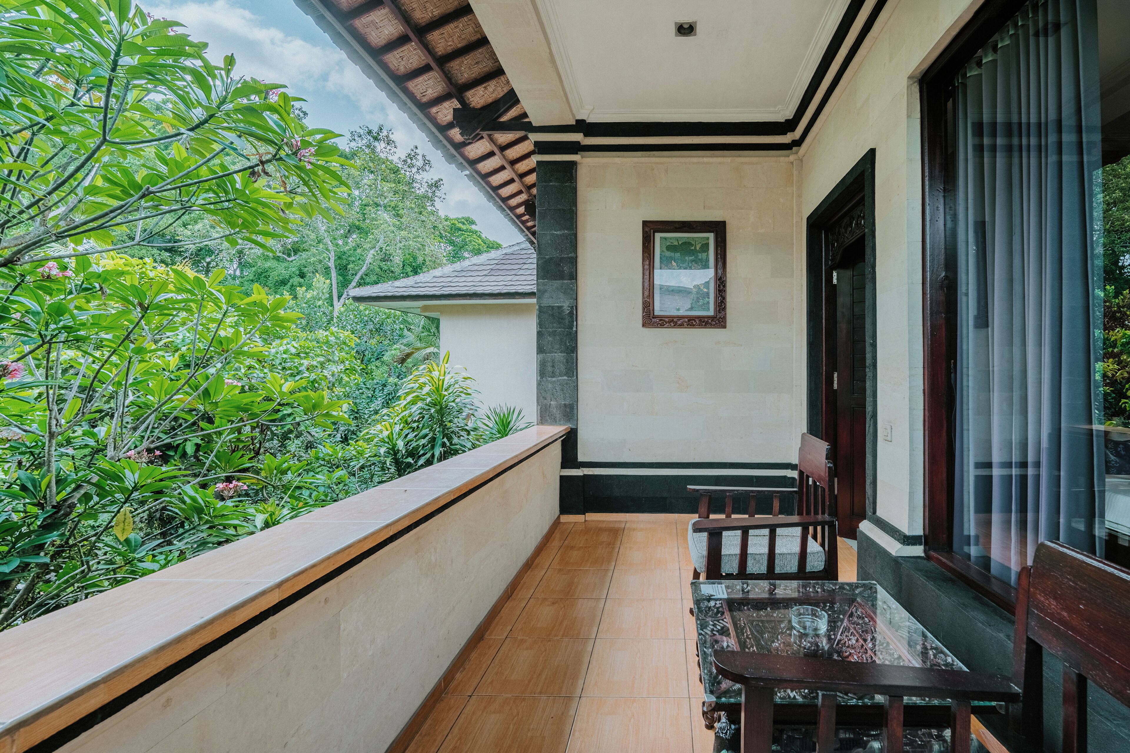 A balcony with a wooden table and chairs, a framed picture on the wall, and a view of greenery.