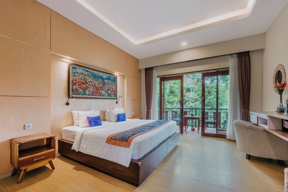 A modern hotel room with a large bed, a desk, and a balcony with a view of greenery.