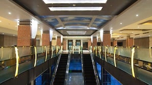 A modern hotel interior with a central escalator flanked by glass railings, a ceiling with a sky mural, and a reflective floor.