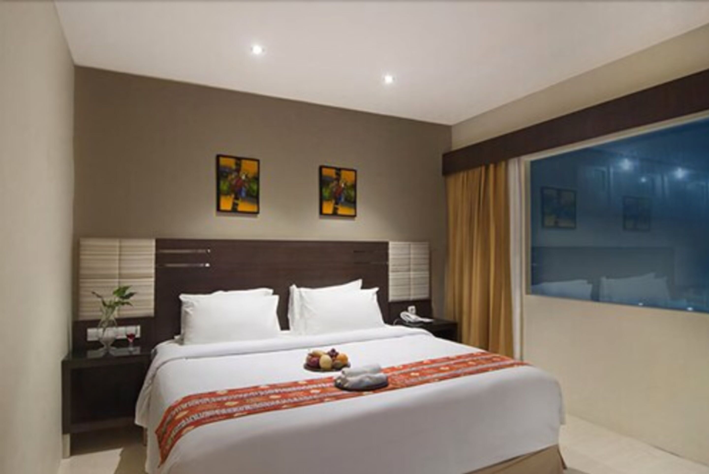 A hotel room with a large bed, two bedside tables, a nightstand, a window with curtains, and two framed pictures on the wall.