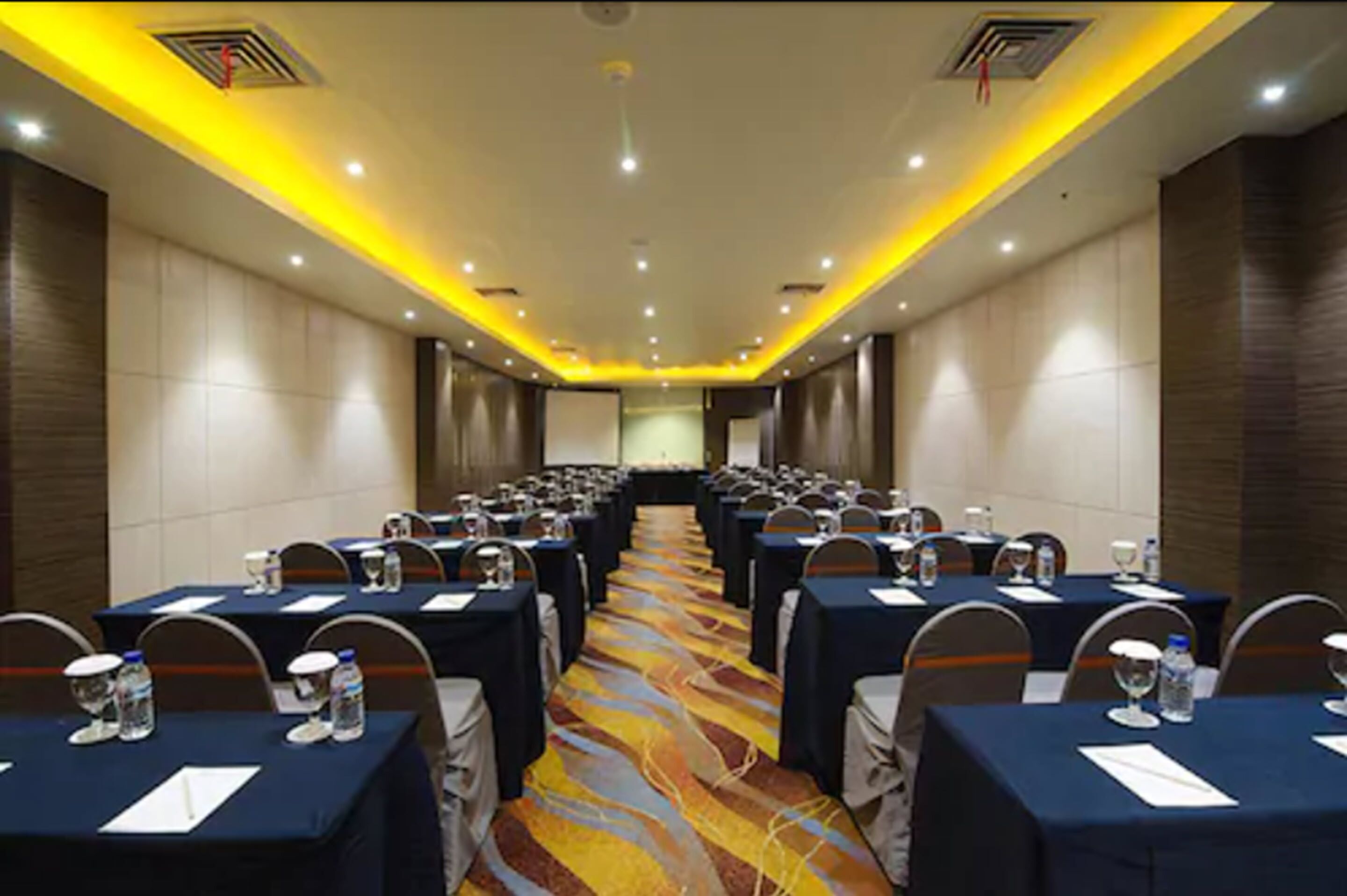 A conference room with rows of tables, each with water bottles and glasses, and a carpeted floor.