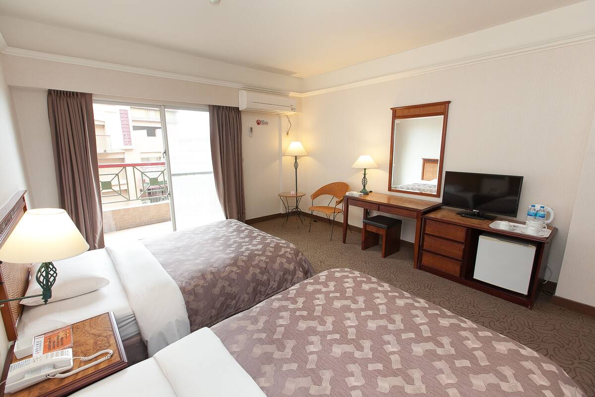A hotel room with two beds, a desk with a television, a small refrigerator, and a window with curtains.