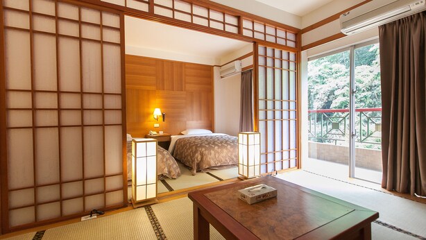 A traditional Japanese-style room with tatami flooring, a sliding shoji screen, a bed with a wooden headboard, a bedside table with a lamp, and a view of a garden through a sliding door. - Hoya Hot Springs Hotel (Taimali)