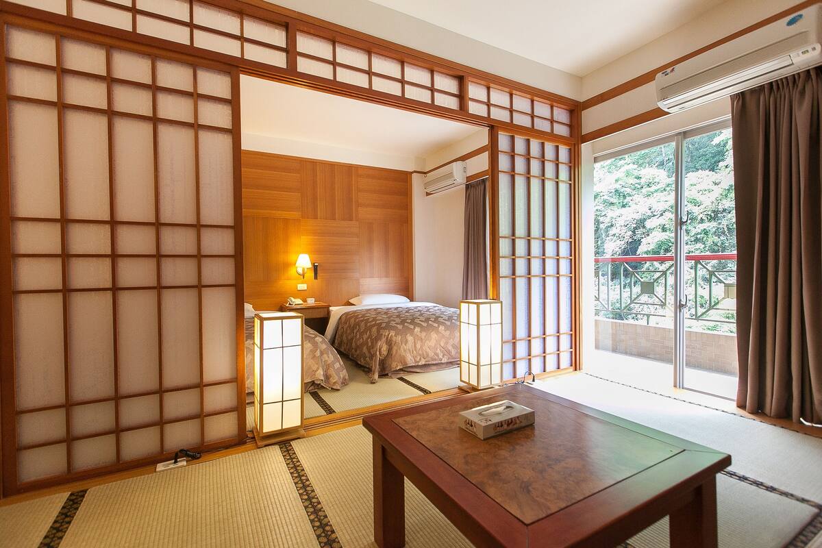A traditional Japanese-style room with tatami flooring, a sliding shoji screen, a bed with a wooden headboard, a bedside table with a lamp, and a view of a garden through a sliding door.