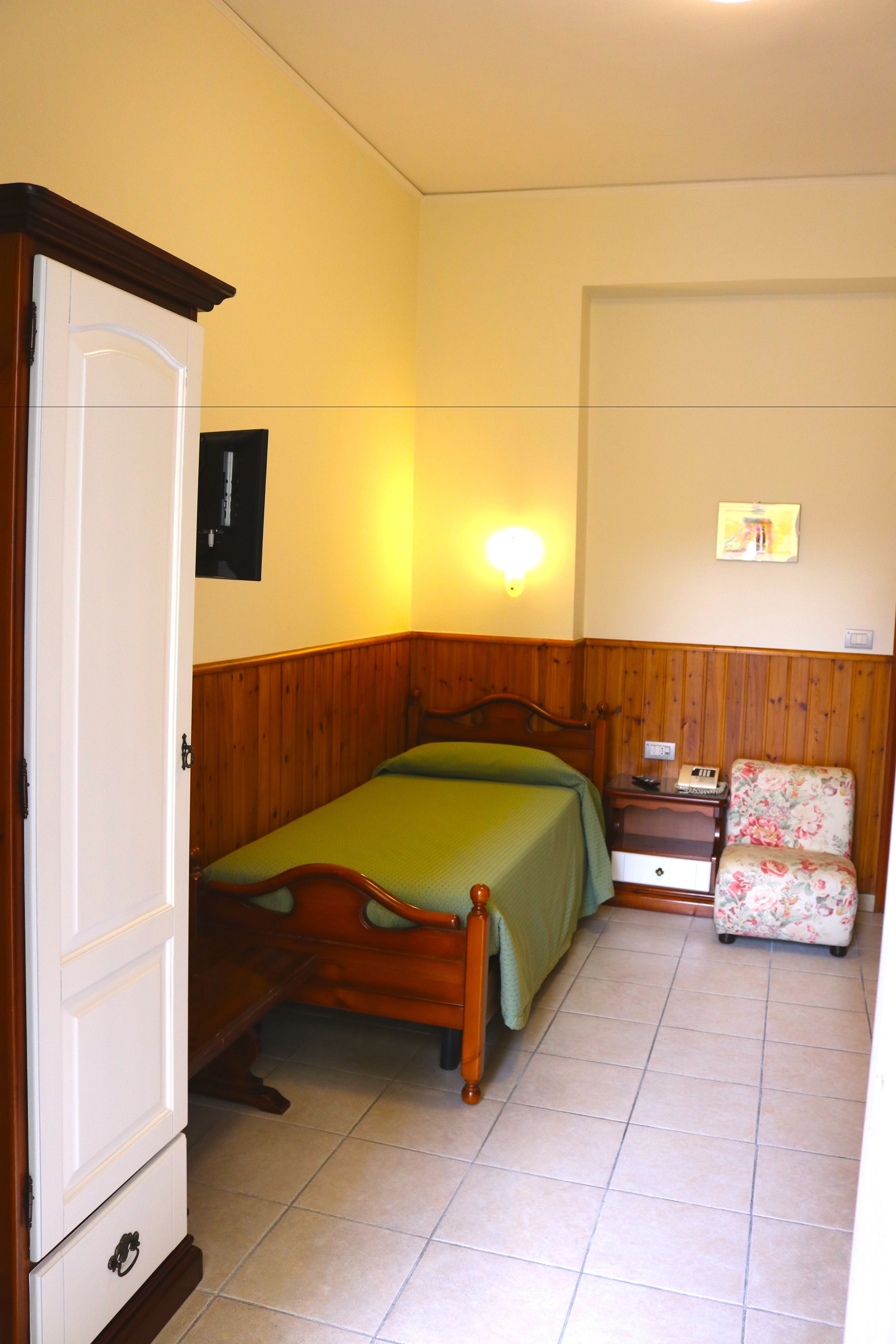 Um quarto com uma cama de madeira, uma mesa de cabeceira, uma cadeira e um candeeiro fixado na parede.