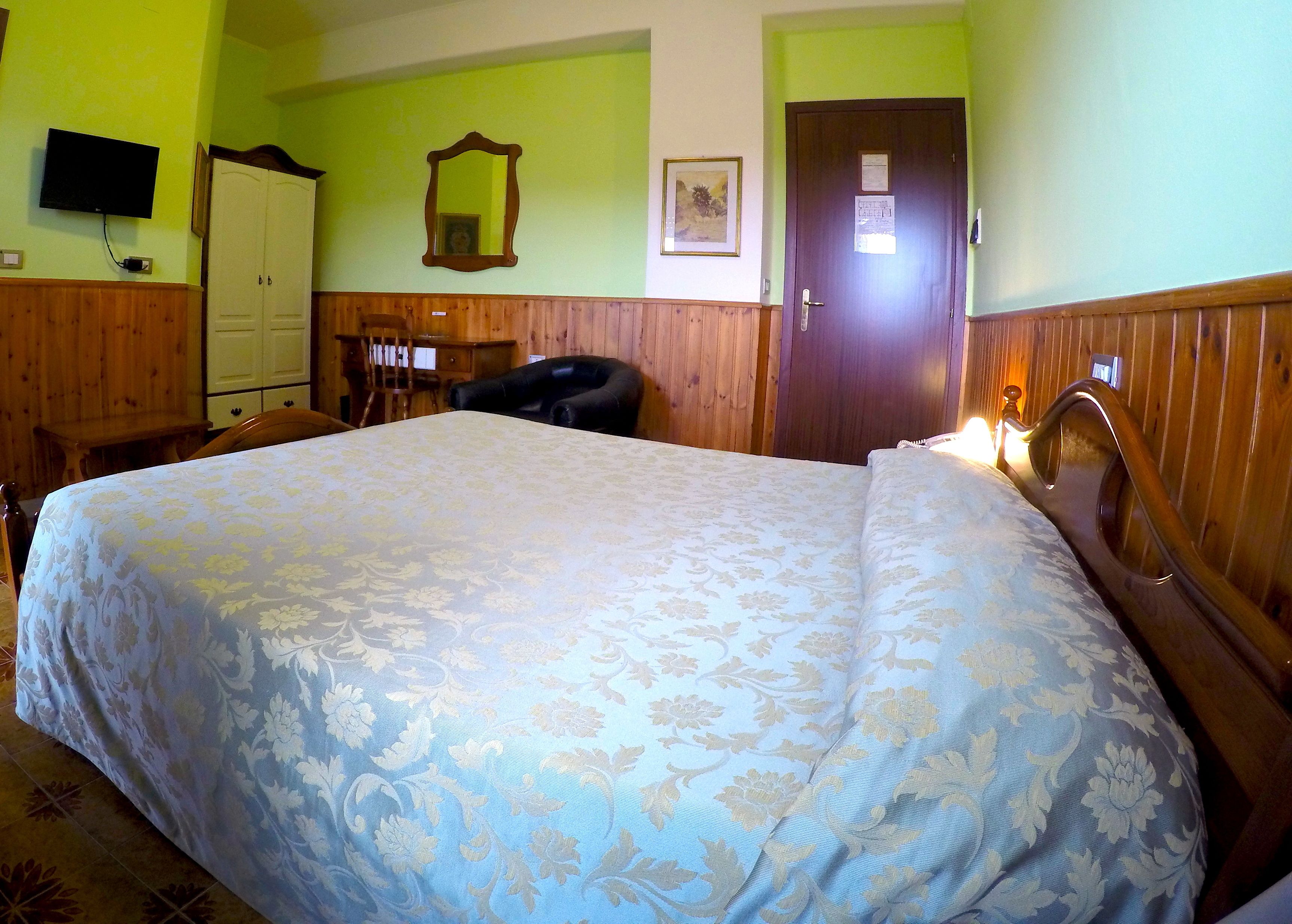 A room with a large bed, wooden headboard, a television on the wall, a door with a notice, and a mirror.
