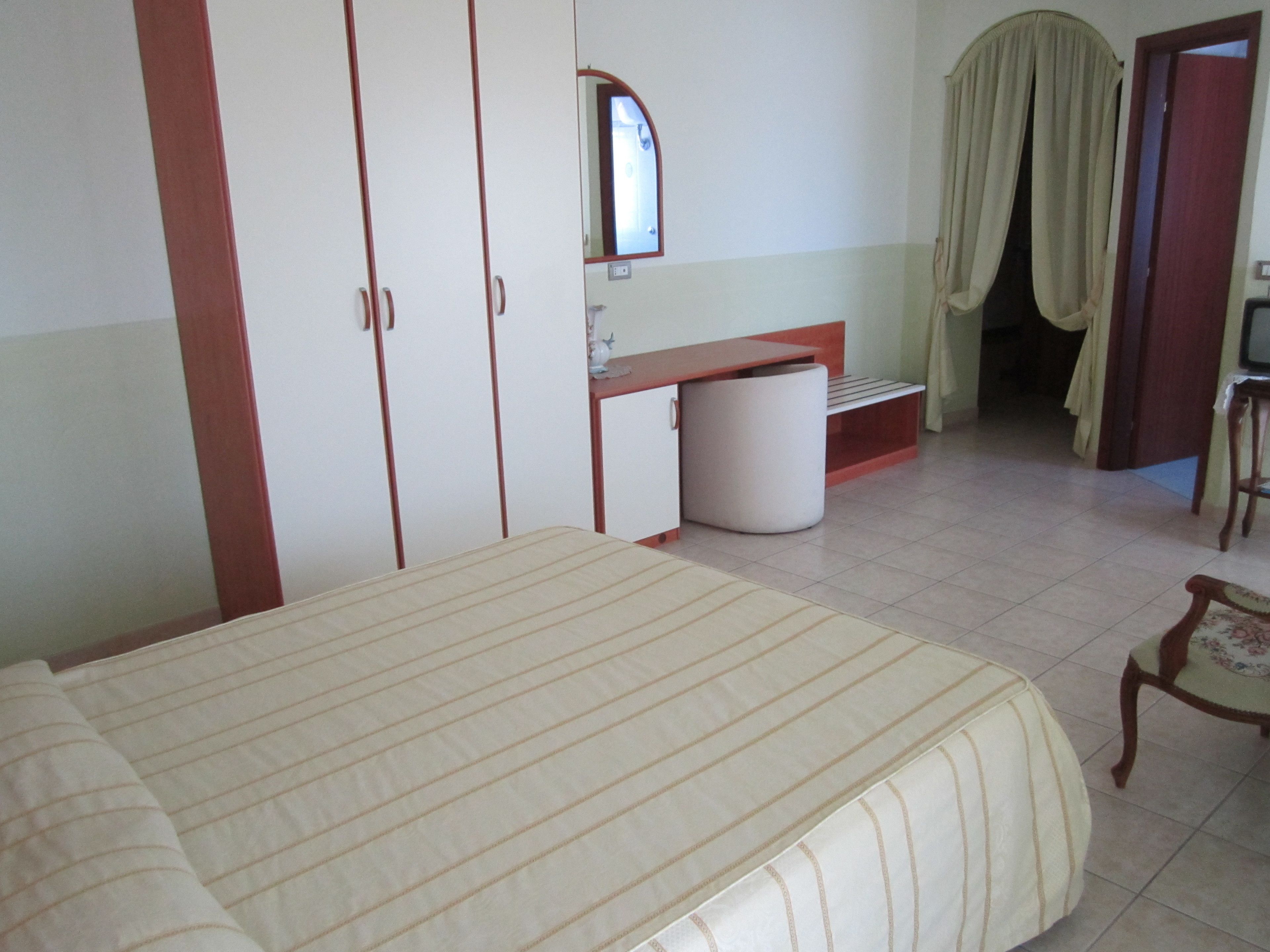 Quarto com cama, guarda-roupa, escrivaninha, cadeira e banheiro visível por uma porta aberta.