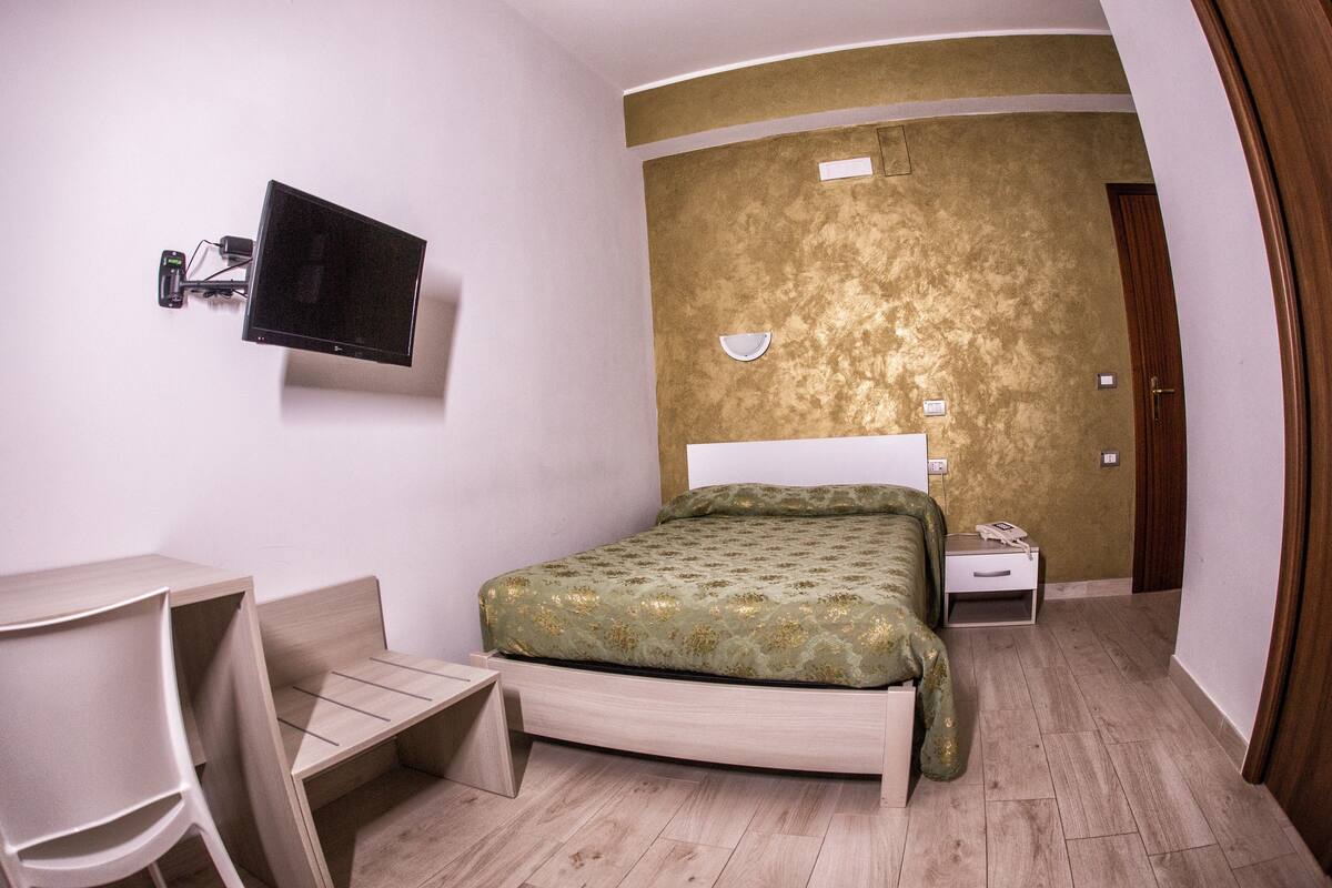 A hotel room with a bed, a desk, a chair, a television, and a wall-mounted light fixture.