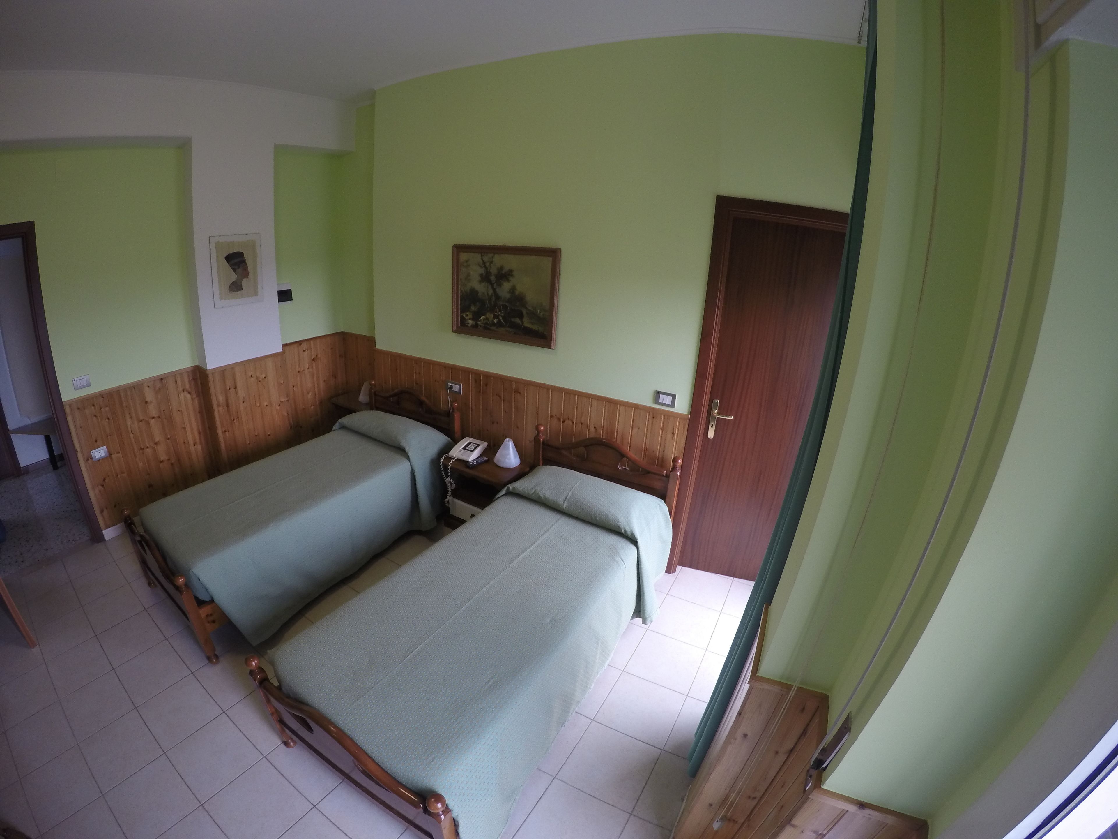 Una stanza con due letti, una testiera in legno, un comodino con una lampada, un quadro appeso al muro e una porta.