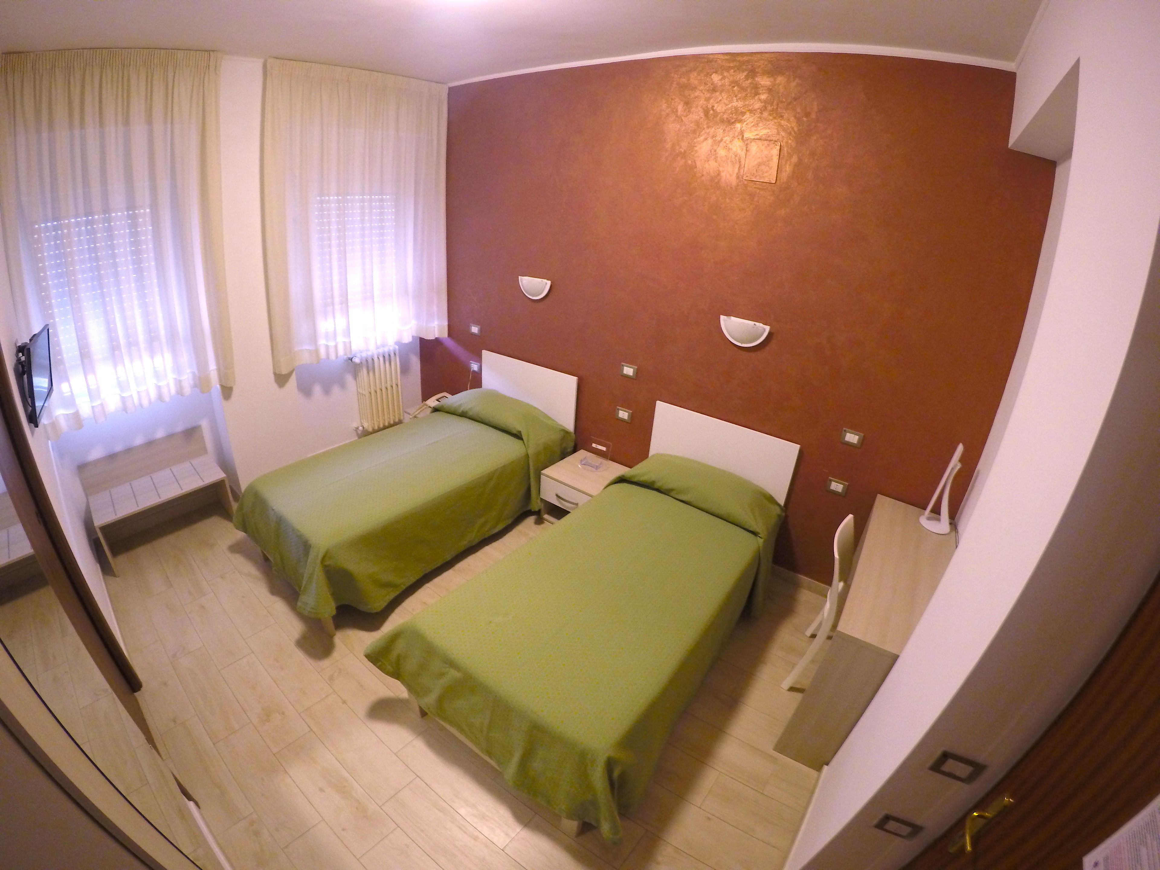Hotellihuone, jossa on kaksi erillistä sänkyä, pieni yöpöytä, tuoli, ikkuna, jossa on verhot, ja seinään kiinnitetty valaisin.
