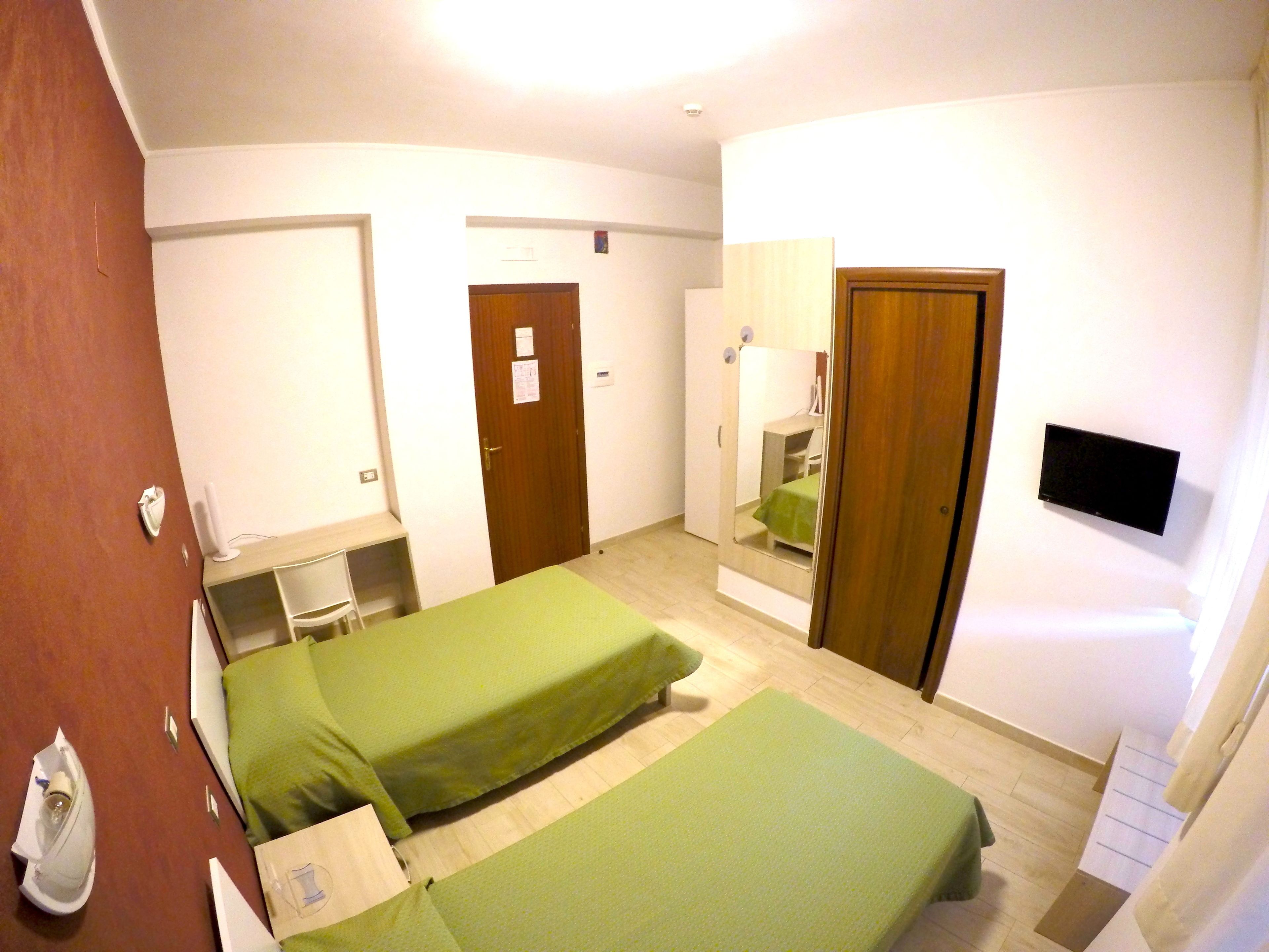 Habitación de hotel con dos camas individuales, un escritorio con silla, un espejo, un televisor montado en la pared y una puerta de madera.
