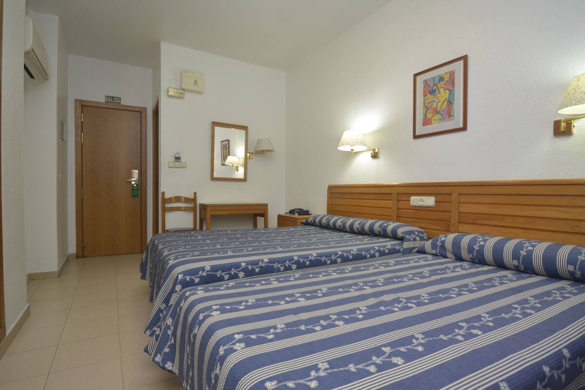 A hotel room with two beds, a wooden headboard, a desk, a chair, a mirror, and wall-mounted lamps.