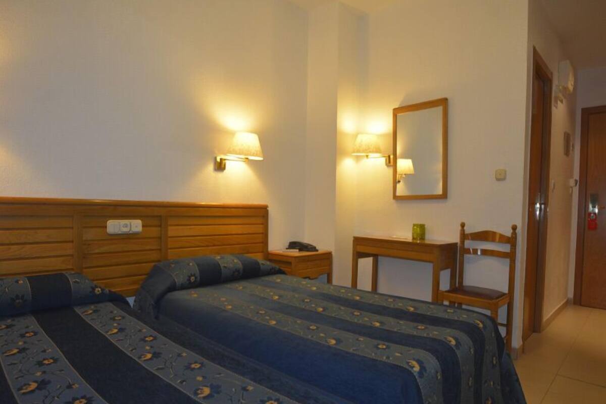 A hotel room with two beds, a bedside table with a lamp, a mirror, and a chair.
