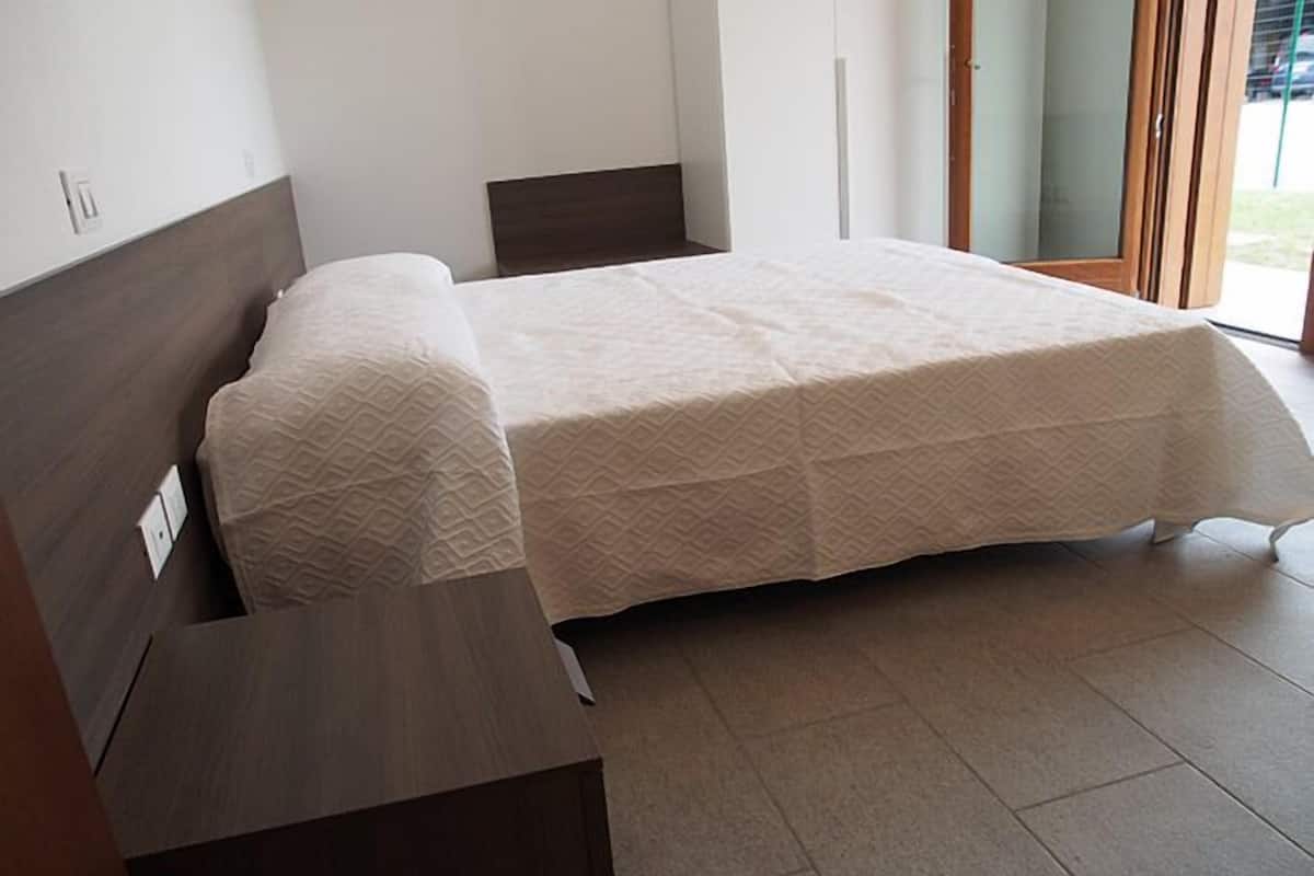 Quarto com cama, mesa de cabeceira e porta para uma varanda.