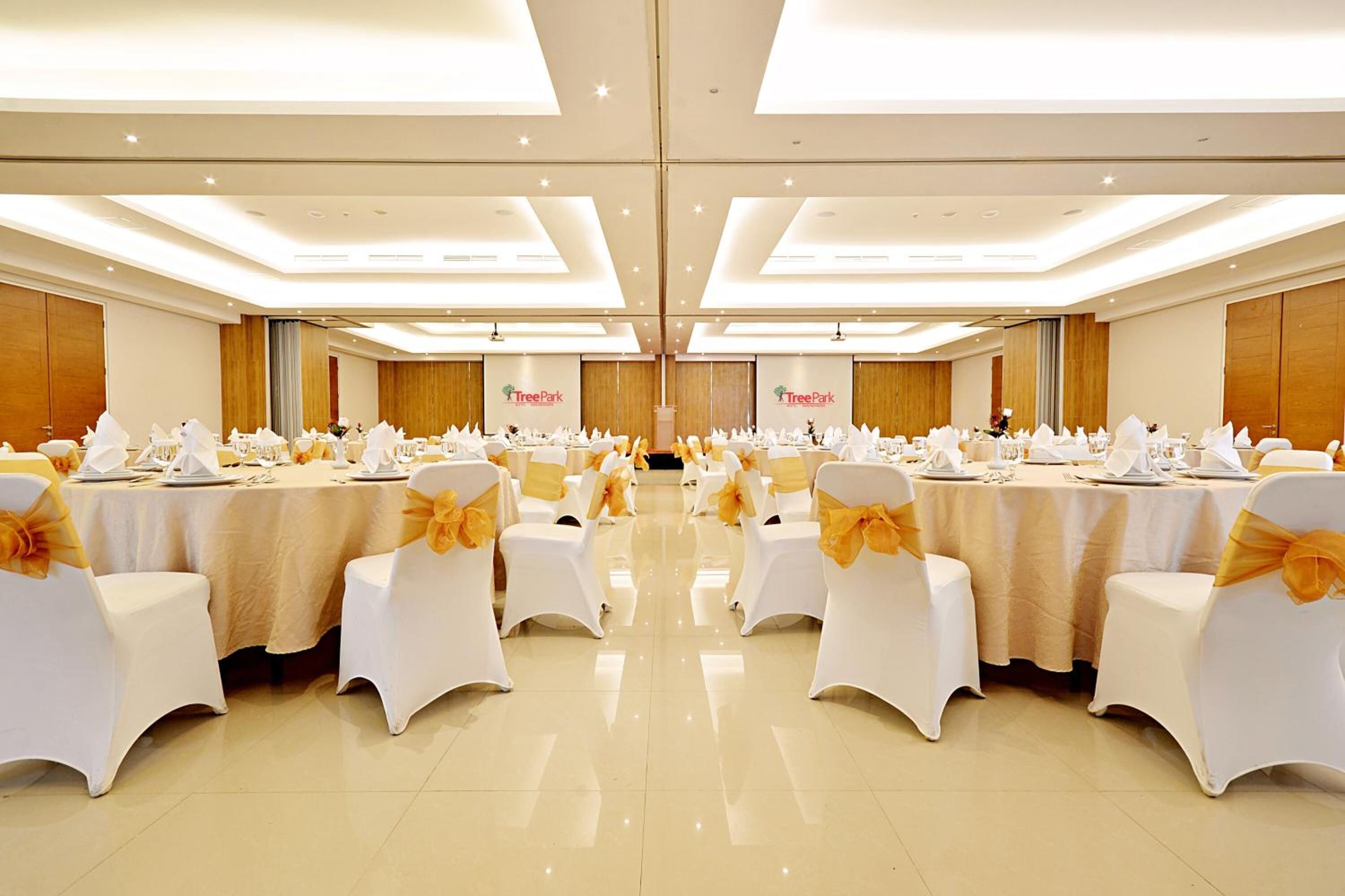 A banquet hall with multiple round tables, each set with white tablecloths and chairs adorned with yellow sashes. The room features a high ceiling with recessed lighting and wooden paneling on the walls.