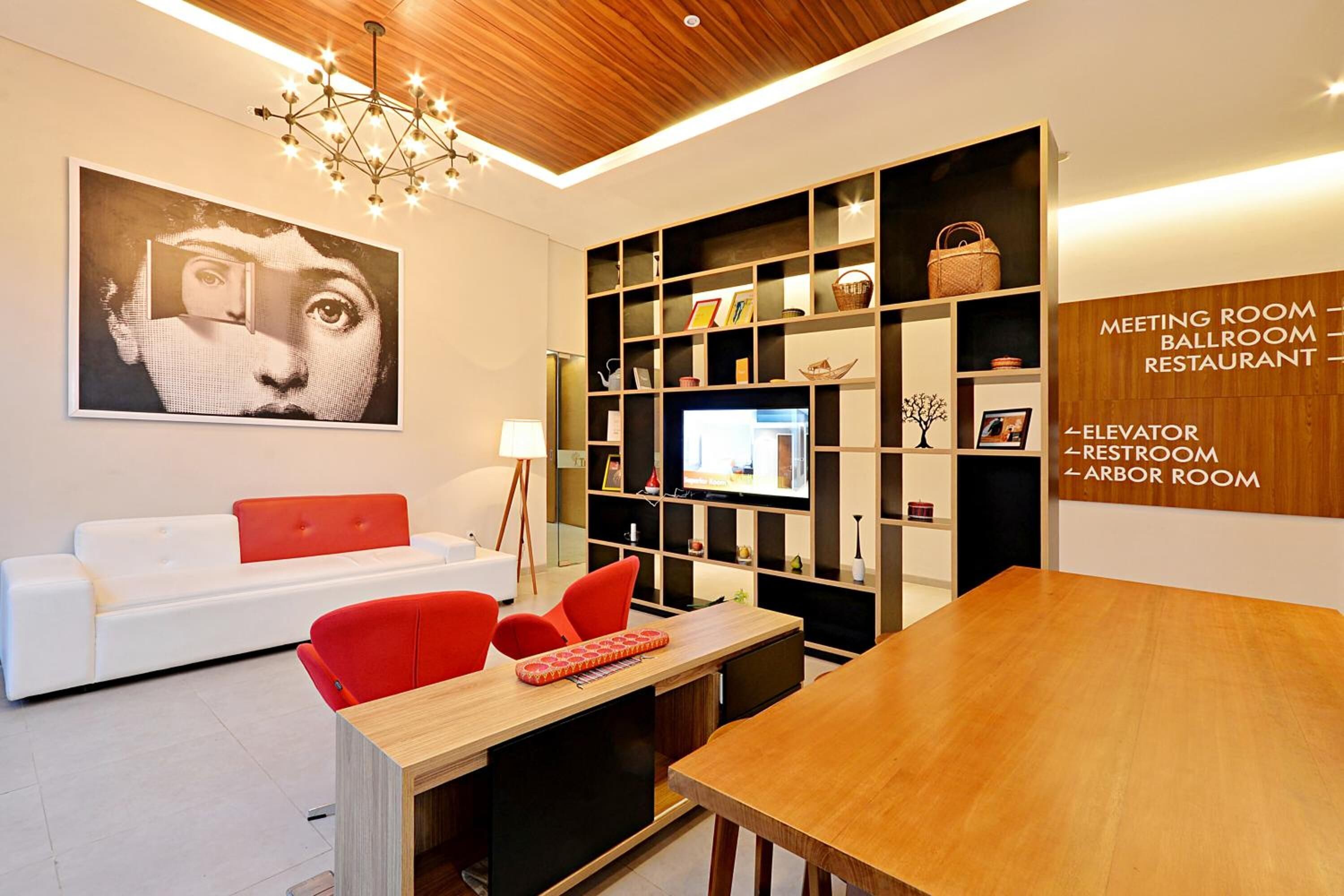 A modern interior with a white sofa, red chairs, a wooden dining table, and a wall-mounted TV. There are shelves with various items and a chandelier.