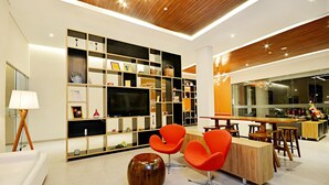 Een moderne woonkamer met een houten plafond, een grote flatscreen televisie, oranje fauteuils, een houten salontafel en een boekenkast met diverse spullen.