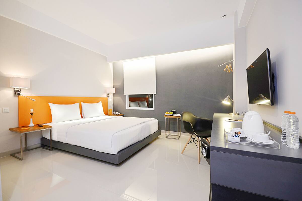 Una habitación de hotel moderna con una cama grande, un escritorio con una silla, una mesita con una lámpara y un televisor de pantalla plana montado en la pared.