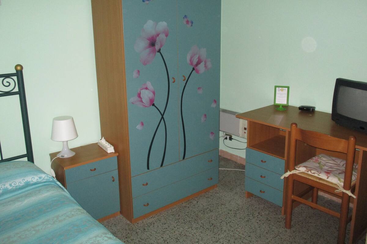 Une chambre à coucher comprenant un lit, un bureau avec un ordinateur, une chaise, une armoire à motifs floraux et une lampe de chevet.