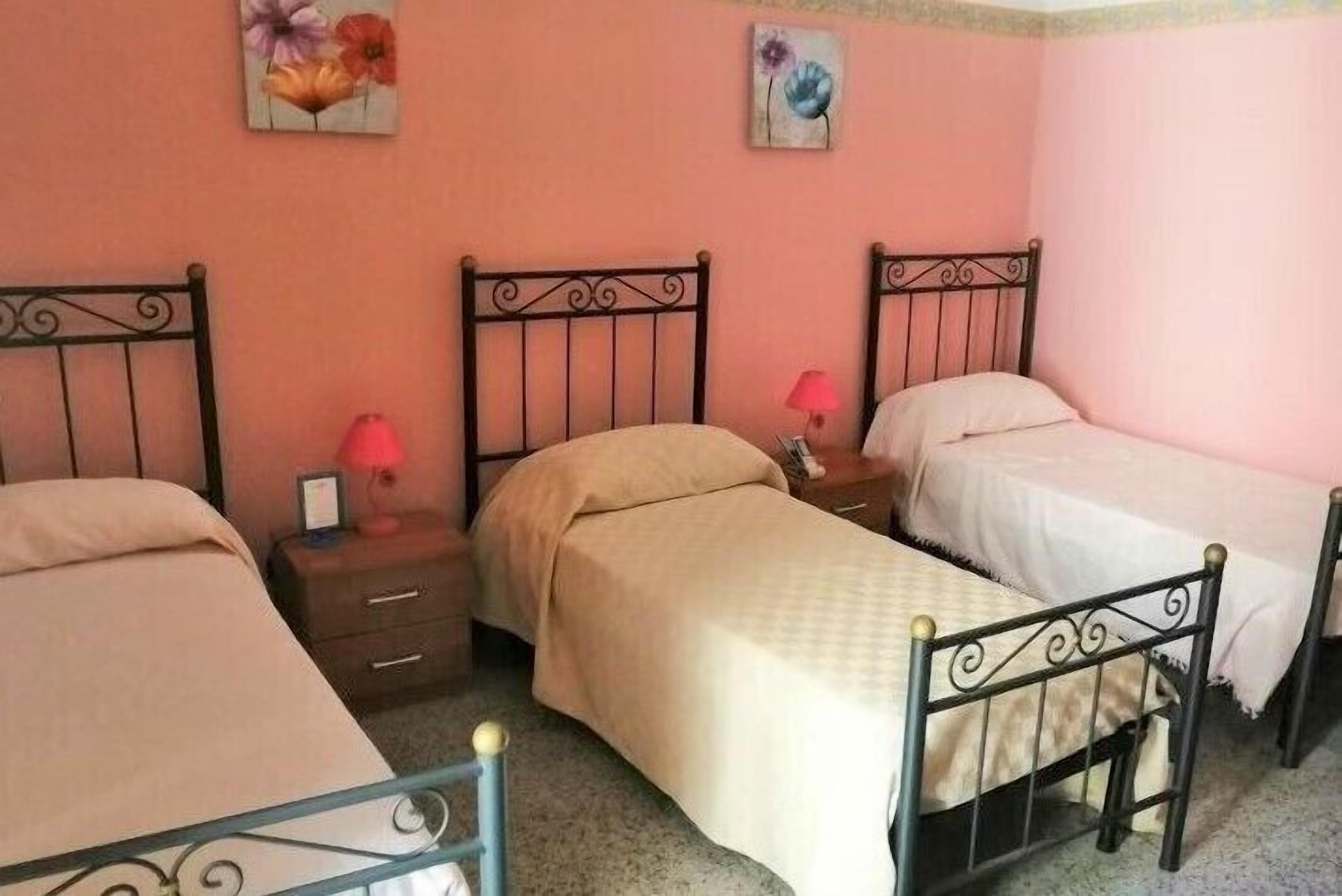 Quarto com três camas individuais, cada uma com estrutura de metal e colchão branco. Há duas mesas de cabeceira, com abajures e uma moldura de foto em uma delas. As paredes são pintadas de rosa, e duas pinturas florais decoram a parede acima das camas.