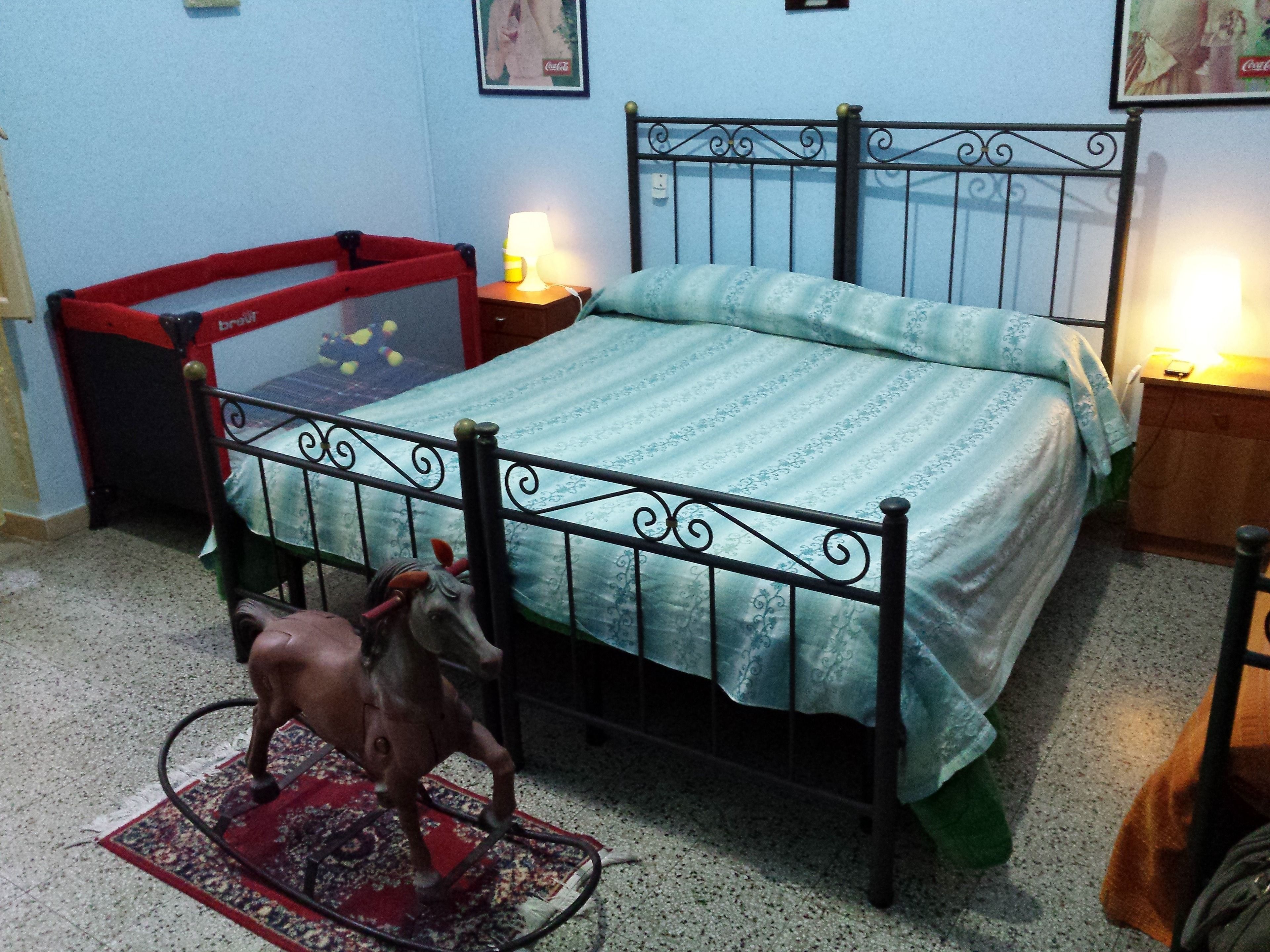 A bedroom with a metal bed frame, a green patterned bedspread, a red chair, a small table with a lamp, and a decorative horse figure on a rug.