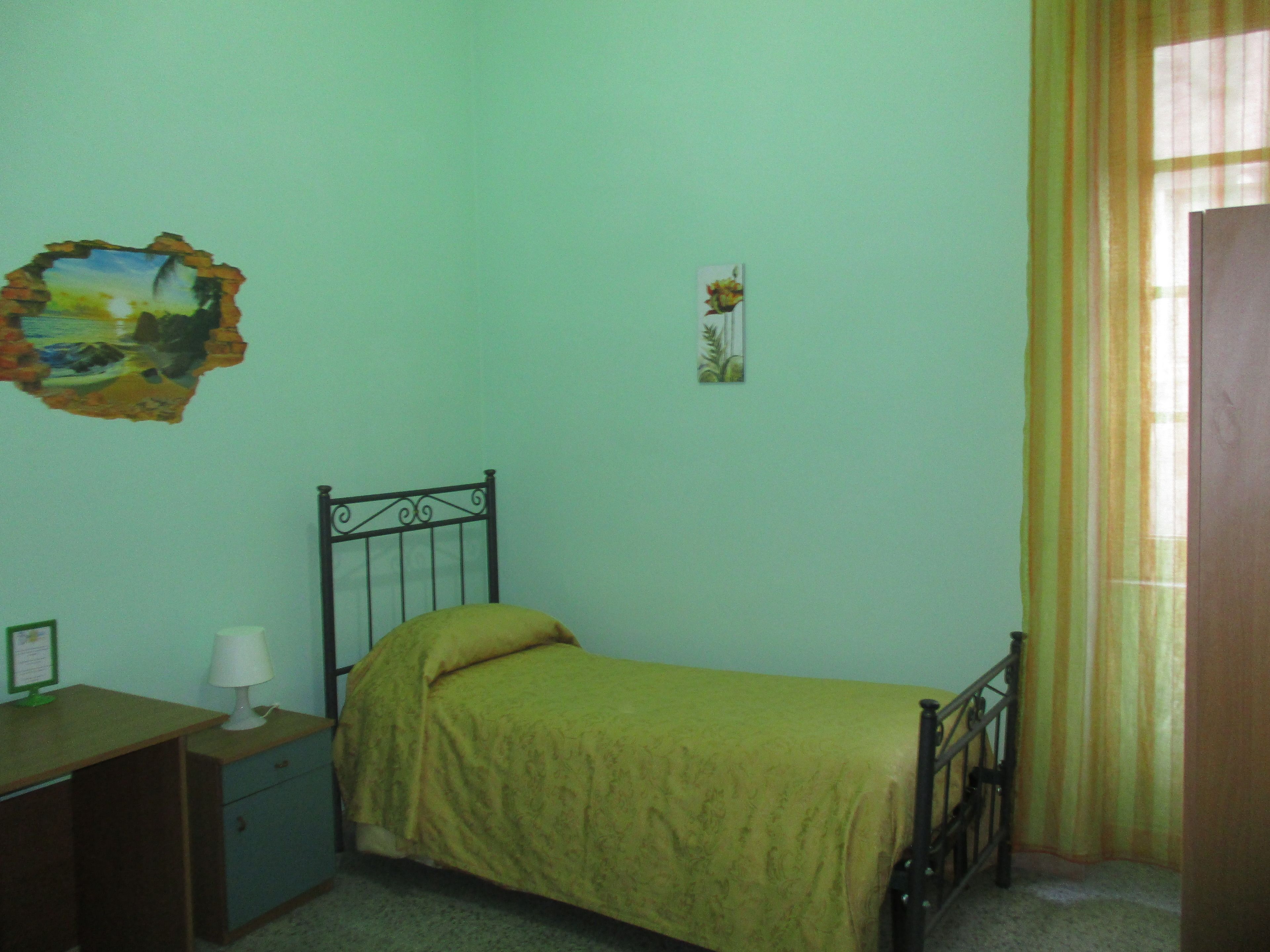 A bedroom with a single bed, a nightstand, a lamp, a small desk, and a window with curtains.