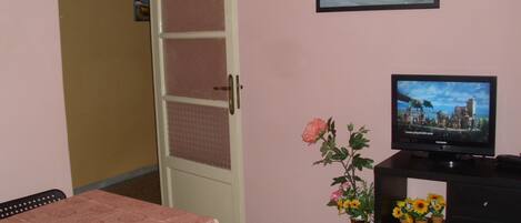A room with a pink wall, a television displaying a game, a table with a pink tablecloth, a chair, a vase with flowers, and a door with a frosted glass panel.