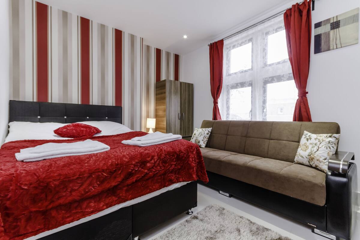 A hotel room with a bed, a sofa, a window with curtains, a lamp, and a painting on the wall.
