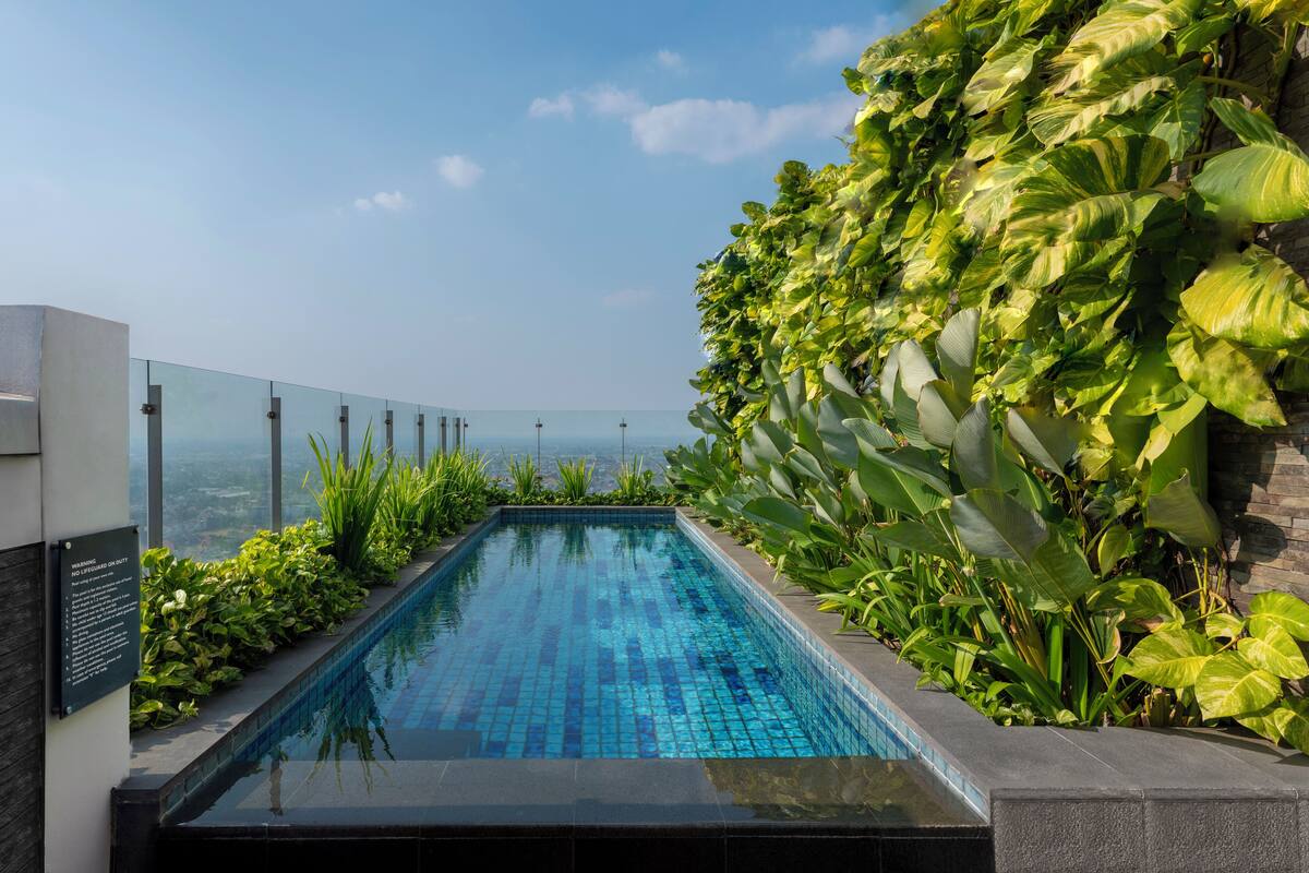 A rectangular infinity pool with clear blue tiles, surrounded by lush green plants and a glass barrier.