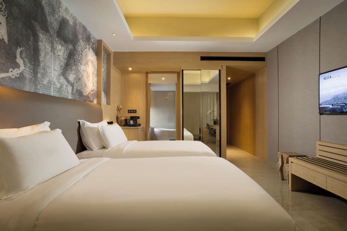 A modern hotel room with two beds, a large artwork on the wall, a television, and a wooden bench.